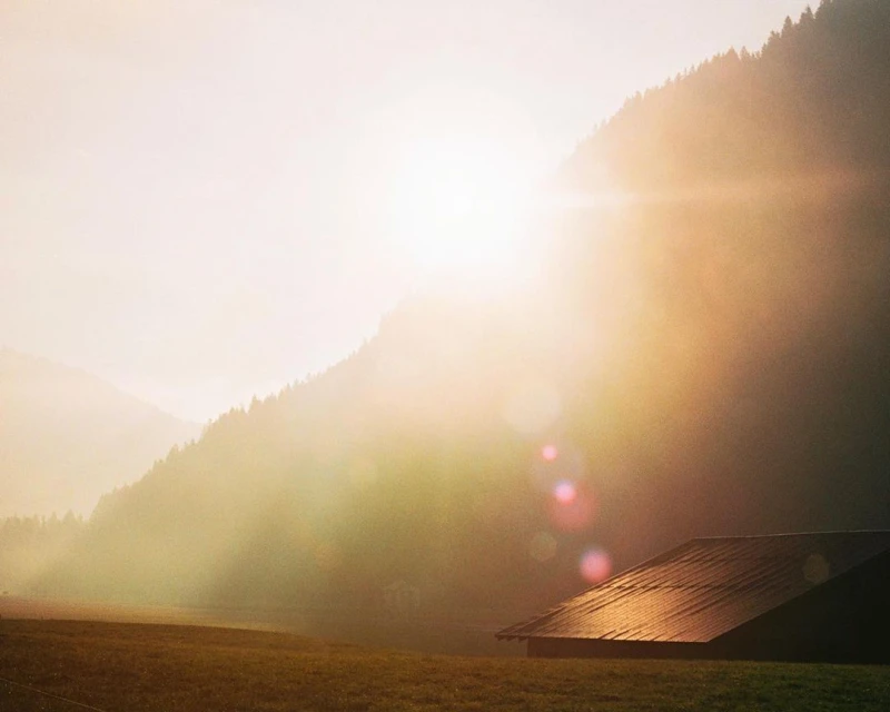 Scenic landscape featuring a sunlit mountainside, a green field, and the roof of a building, creating a serene and natural scene.