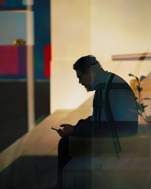 A silhouette of a man wearing a cap and glasses sits using a smartphone, creating a sense of urban quiet and solitary tech use.