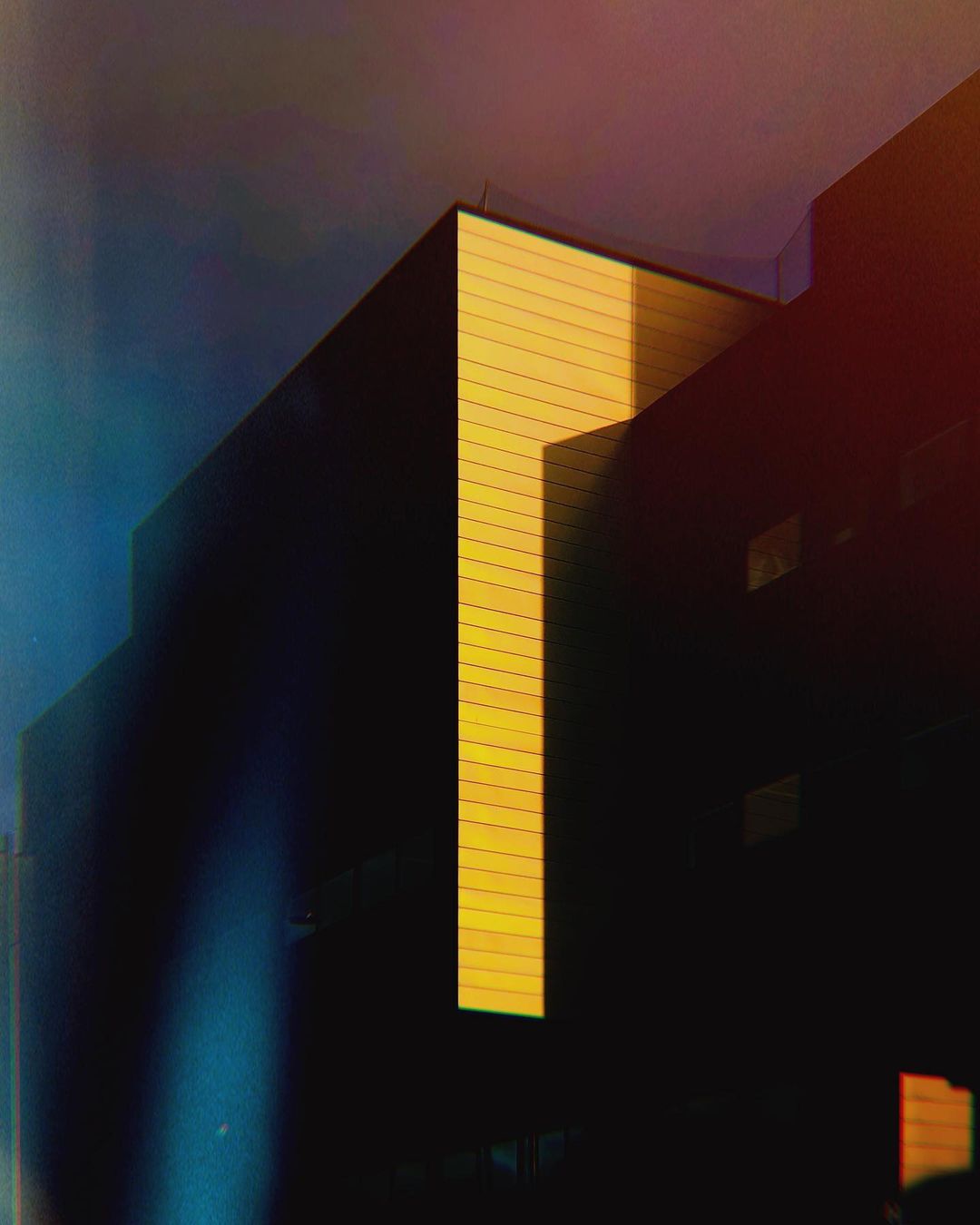 Exterior view of the corner of a dark building with a modern architectural style, featuring a vertical yellow accent.