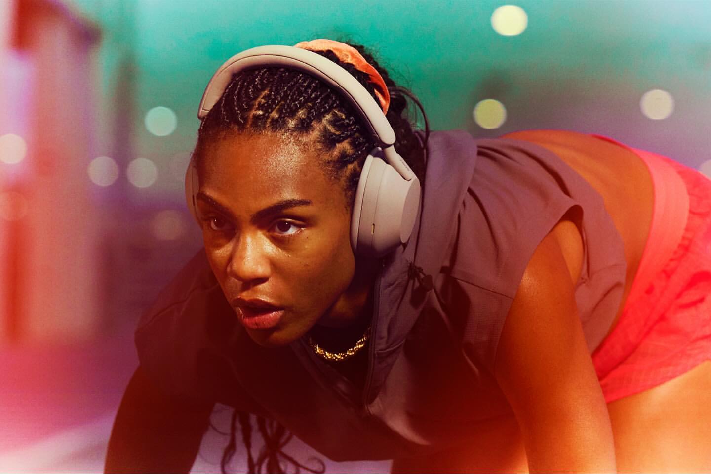 Close-up of a woman with wireless headphones, preparing for a fitness activity in a blurred urban setting with teal and orange hues.