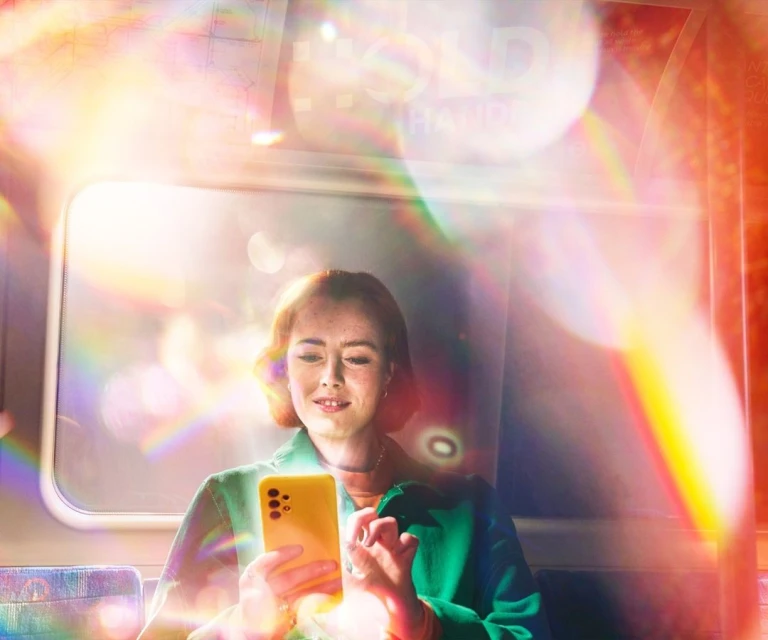 Young woman uses smartphone on subway train, wearing green jacket, with light flares and urban transport environment visible.