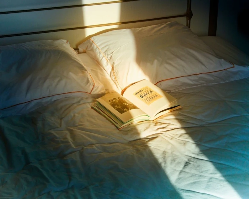 Still life of an open book on a made bed with sunlight casting shadows. Pillows, white sheets, and soft light create a cozy scene.