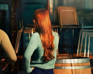 Woman with long red hair wearing a sage green knitted sweater standing indoors with stacked chairs in background