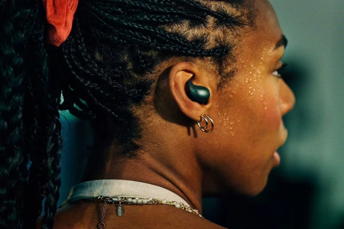Close-up profile of a black woman wearing dark wireless earbuds, gold hoop earrings, and a white top with a beaded necklace.
