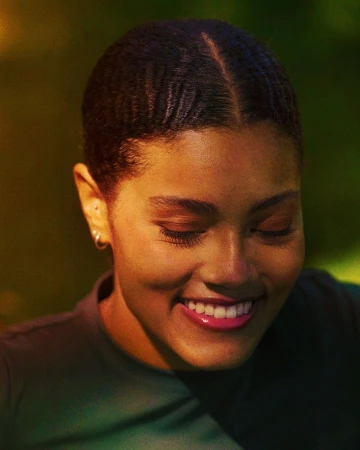 Close-up portrait of a smiling woman with braided dark hair wearing a dark top against a dimly lit background.