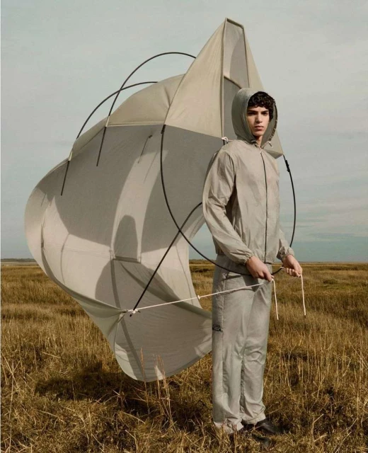 A young adult male in a light grey hooded tracksuit holds a large, deconstructed kite outdoors in a grassy field under a pale blue sky.