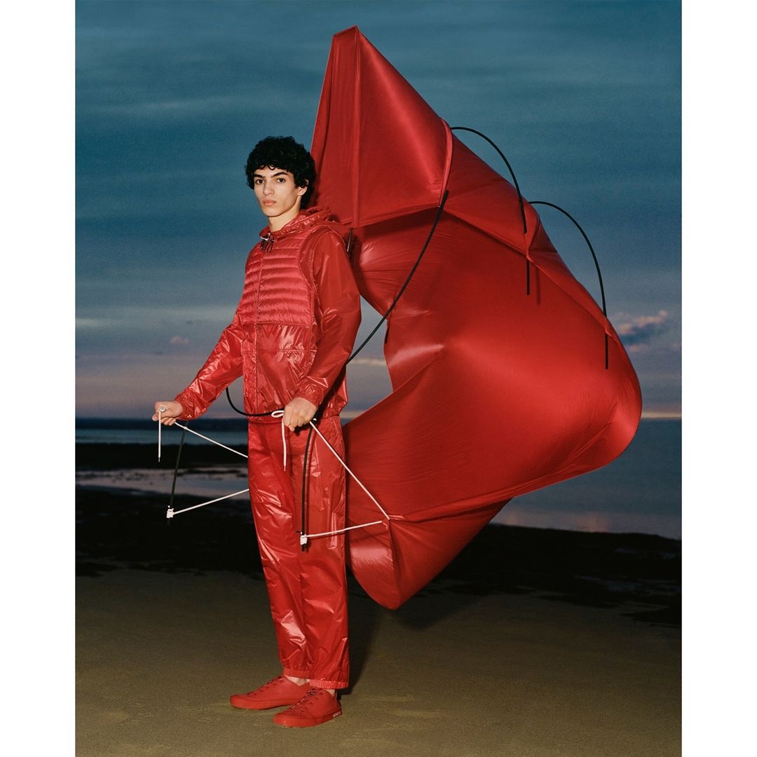 A young adult male stands on a beach holding a large red kite, wearing a matching red puffer vest, tracksuit, and sneakers against a twilight sky.
