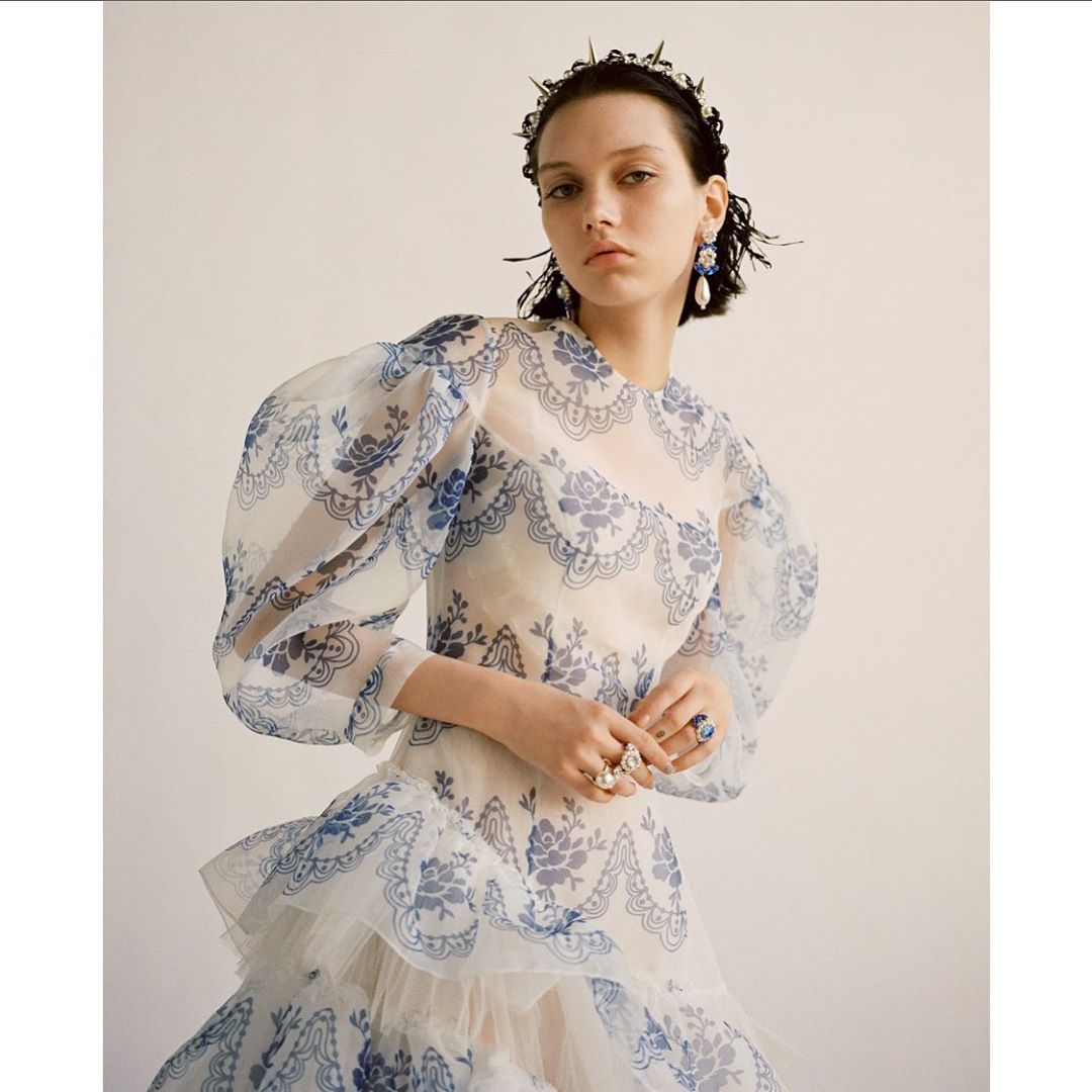 Elegant portrait of a woman in a sheer, patterned dress with statement jewelry against a minimalist background, highlighting fashion and beauty.