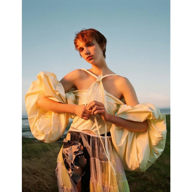 Woman with short red hair poses in a light yellow top with large sleeves and a multicolored skirt against a sky and grassy background.