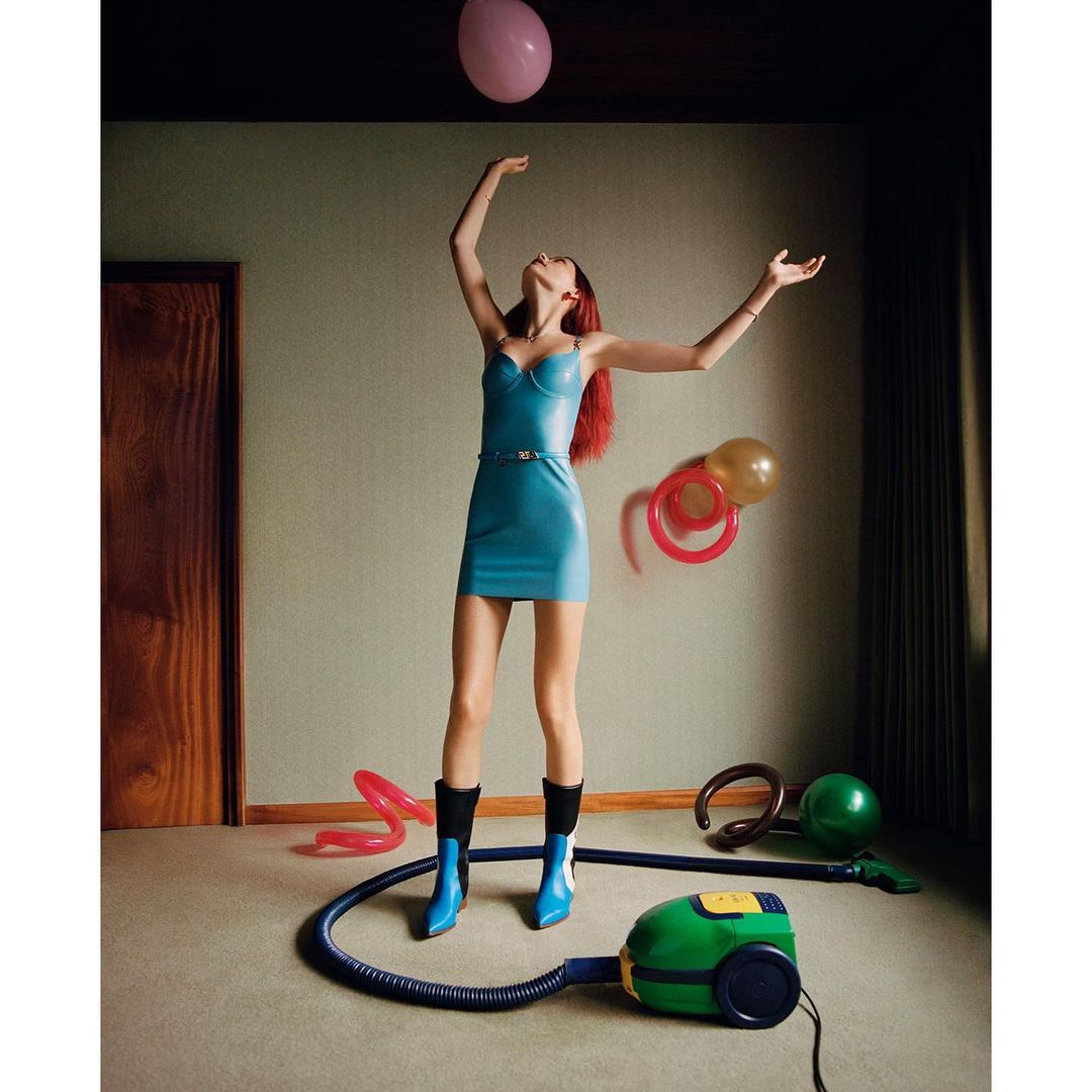 Woman in a blue dress with a balloon indoors, styled fashion shoot, surrounded by other balloons and a green vacuum cleaner.