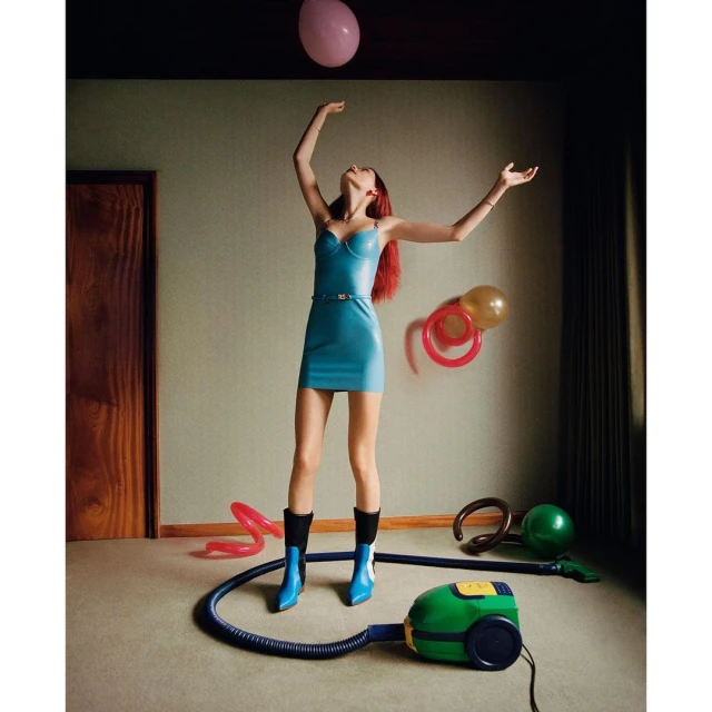 Woman in a blue dress with a balloon indoors, styled fashion shoot, surrounded by other balloons and a green vacuum cleaner.