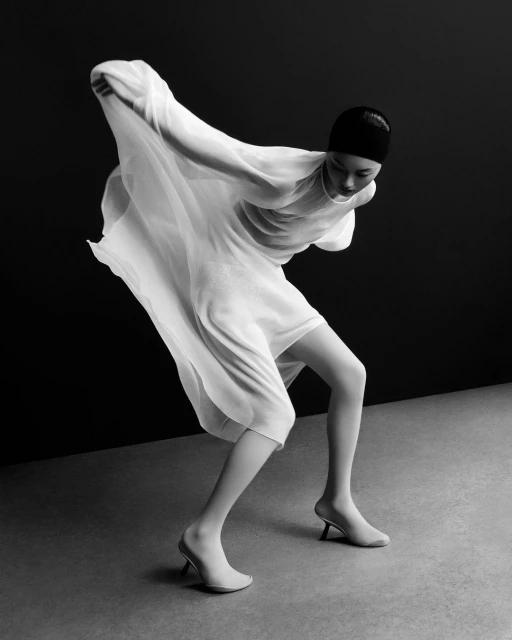 A model poses in a white dress and low heels in a black and white studio fashion shot with minimalist elegance and graceful movement.