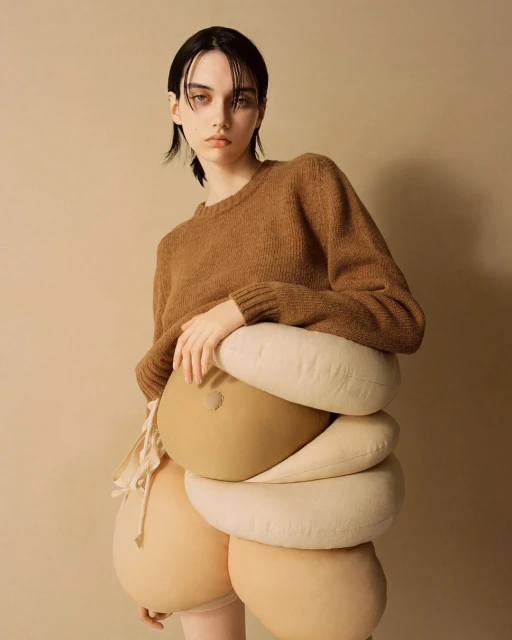 Brunette model poses in a brown sweater with beige, padded costume pieces, in a studio shot on beige background. 