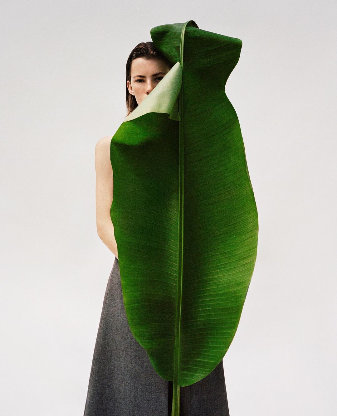 A young woman partially obscured by a large green banana leaf, creating a natural and minimalist fashion portrait.