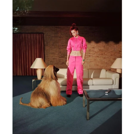 Woman in pink outfit poses indoors with Afghan hound dog. Modern interior features brick walls, sofa, and blue carpet.