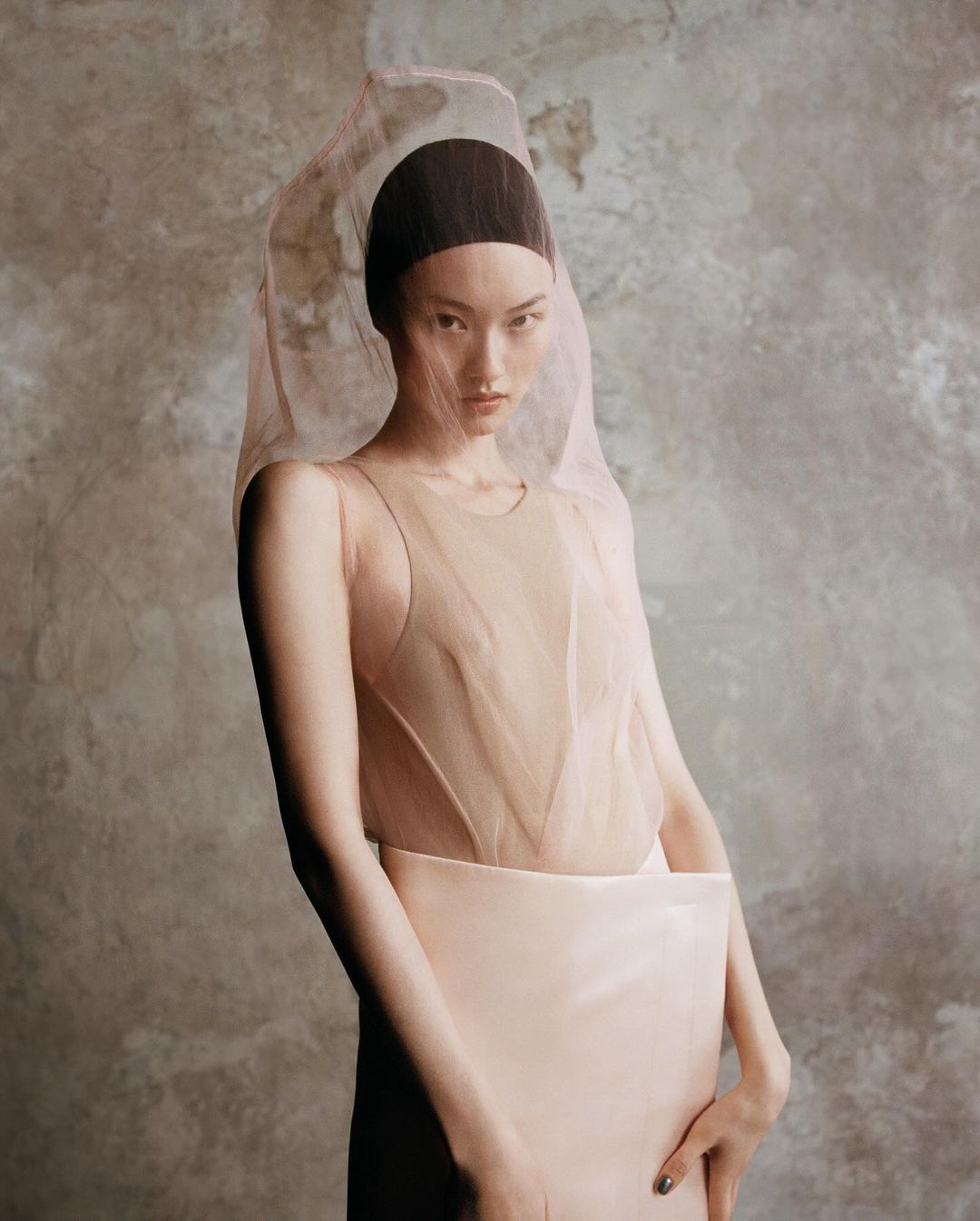 A serene portrait of an Asian woman in a sheer veil, sleeveless top, and a peach skirt, set against a textured neutral background.
