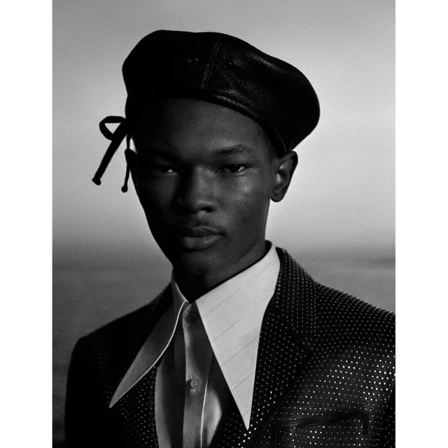 Portrait of a young black man wearing a beret and embellished jacket, set against a light gradient backdrop. Black and white, fashion.