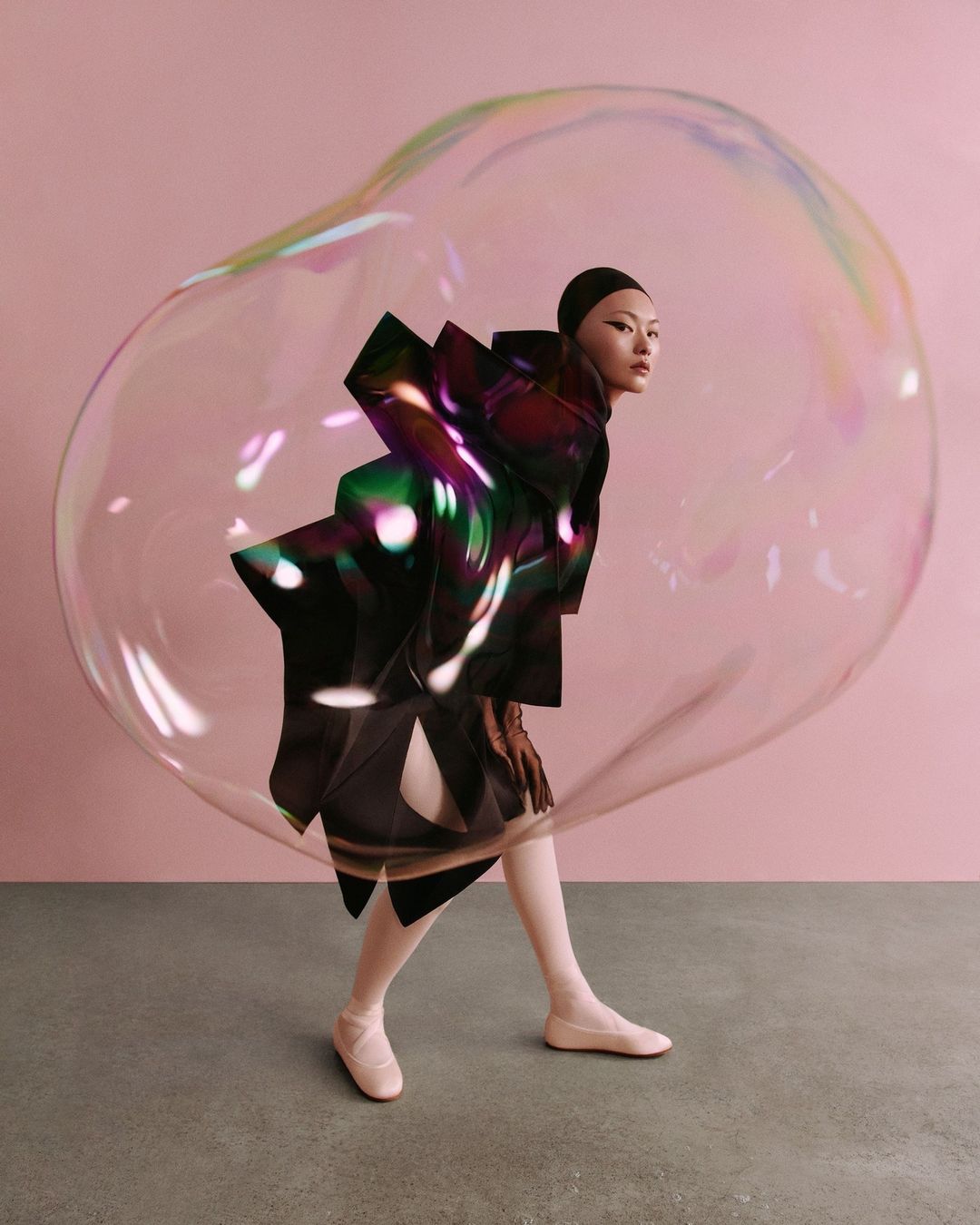 Asian woman in avant-garde fashion poses inside a large iridescent bubble against a pink studio backdrop, showcasing unique style.