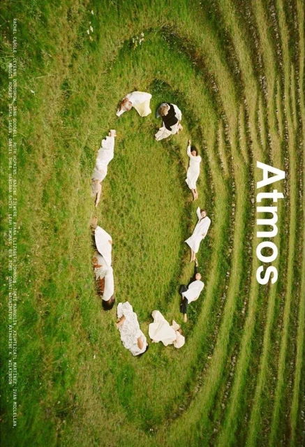 Overhead shot of multiple people in white garments lying in a circle on green grass in an outdoor setting with artistic styling.