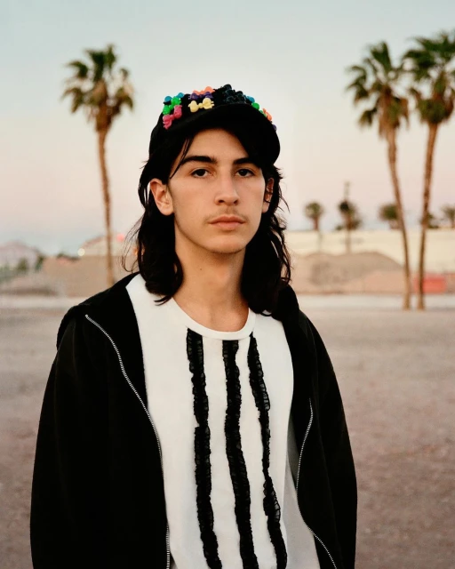 Portrait of a young adult with long dark hair wearing a decorated baseball cap, t-shirt, and jacket in front of palm trees outdoors.