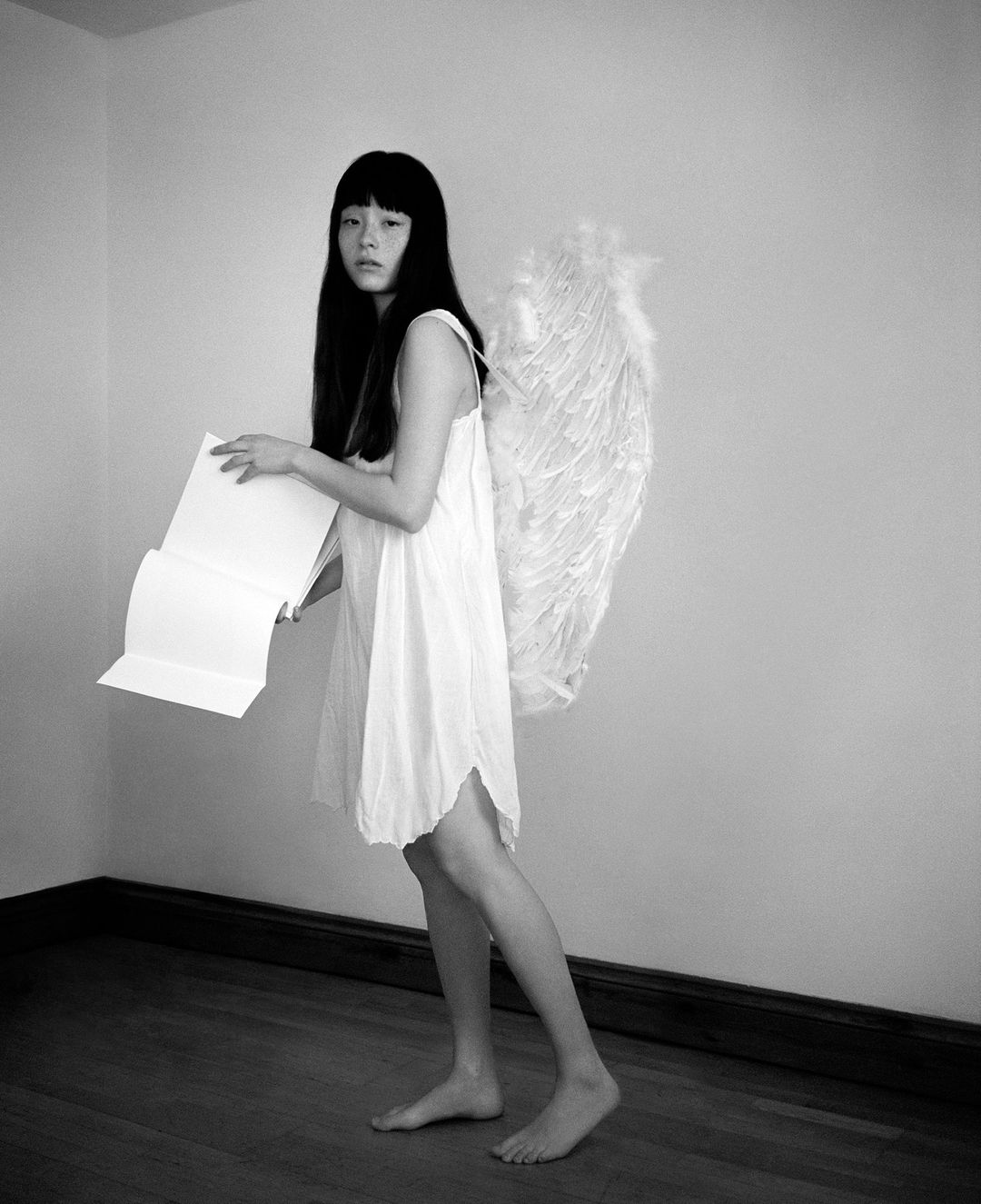 Full shot of a young adult female with dark hair and feather wings in a white dress holding a paper in an indoor setting against a wall.
