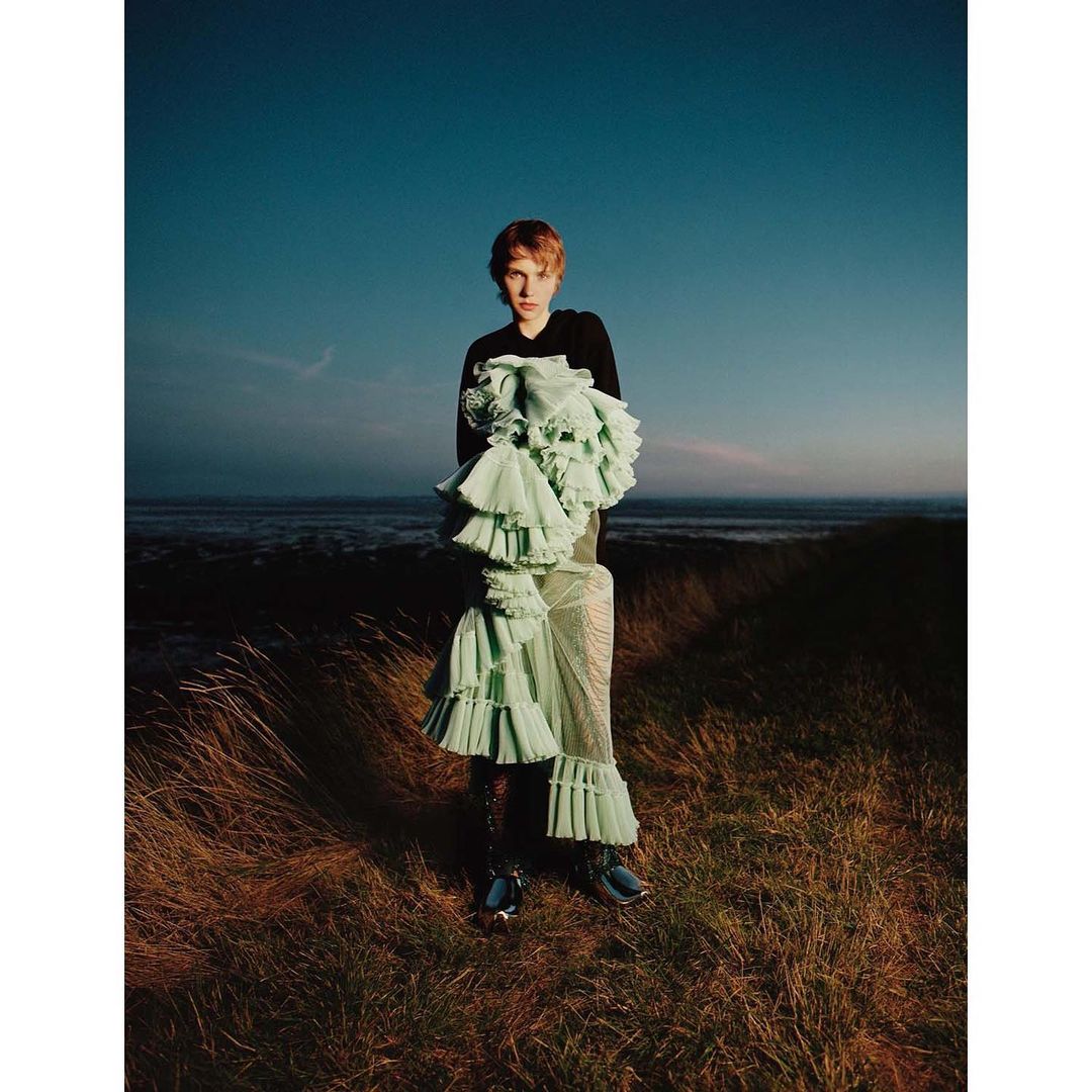 A person stands in a field near the ocean, wearing a black top and a light green dress with ruffles, in an outdoor fashion editorial.