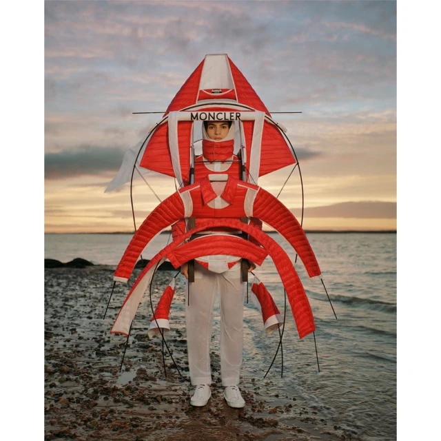 Fashion editorial shot of a person wearing a red and white Moncler outfit on a rocky beach with a colorful sunset sky.