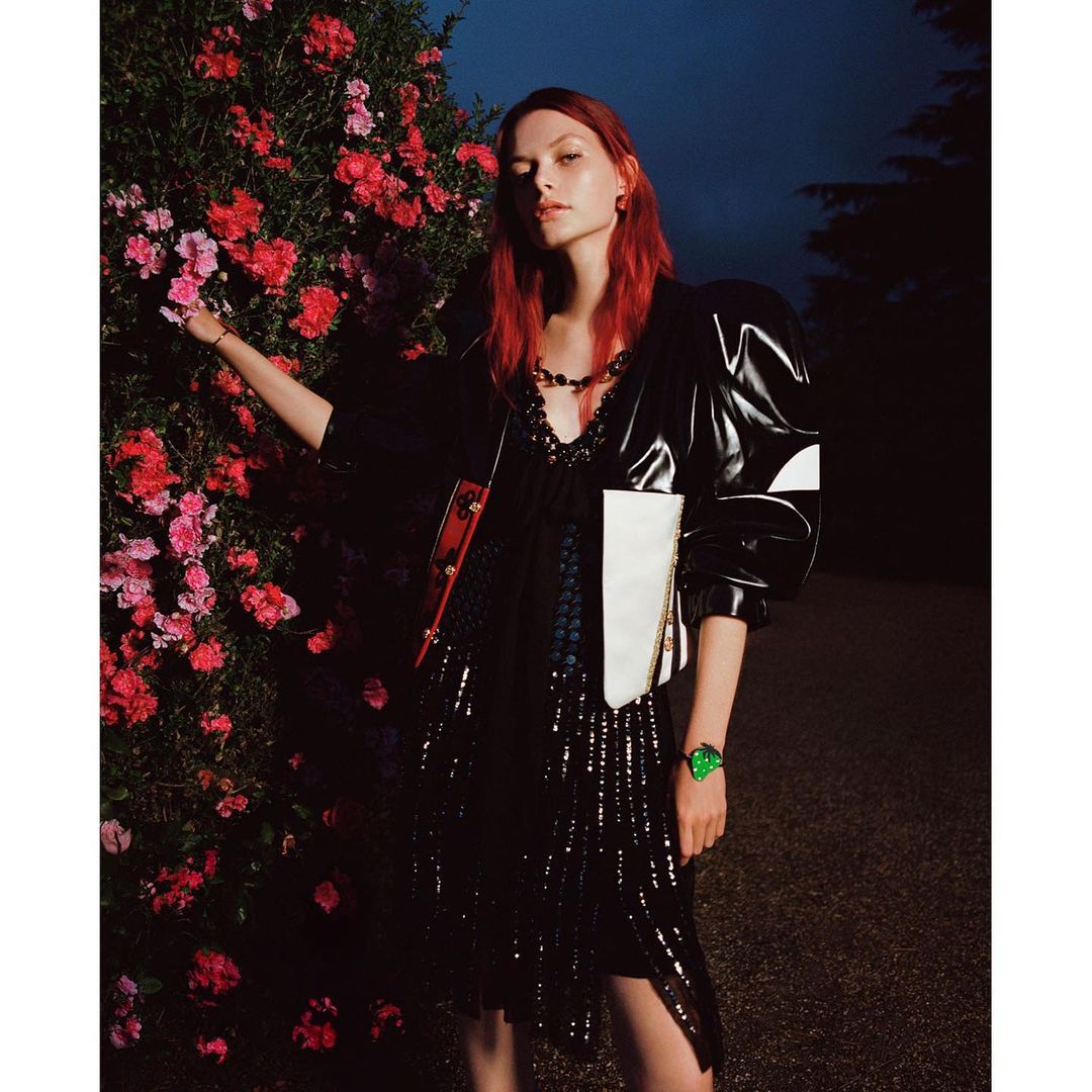 Fashionable woman with red hair poses in a black dress and jacket, standing beside a vibrant pink flower bush at night.