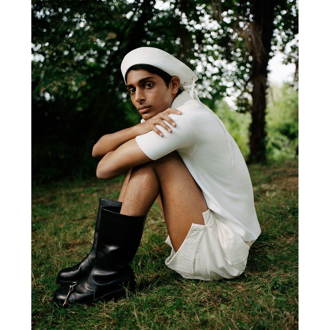 Young adult sits on grass in a white outfit and black boots against a green outdoor background in natural light, fashion editorial.