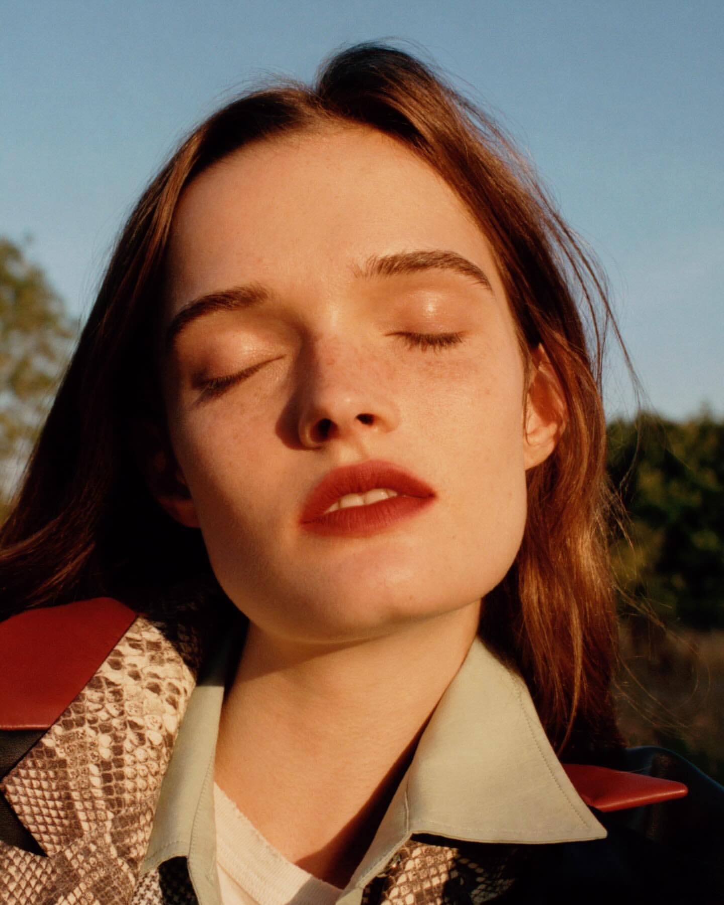 Close-up of a young adult woman with closed eyes wearing a snakeskin jacket. Natural lighting illuminates her face in an outdoor environment.