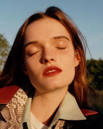 Close-up of a young adult woman with closed eyes wearing a snakeskin jacket. Natural lighting illuminates her face in an outdoor environment.