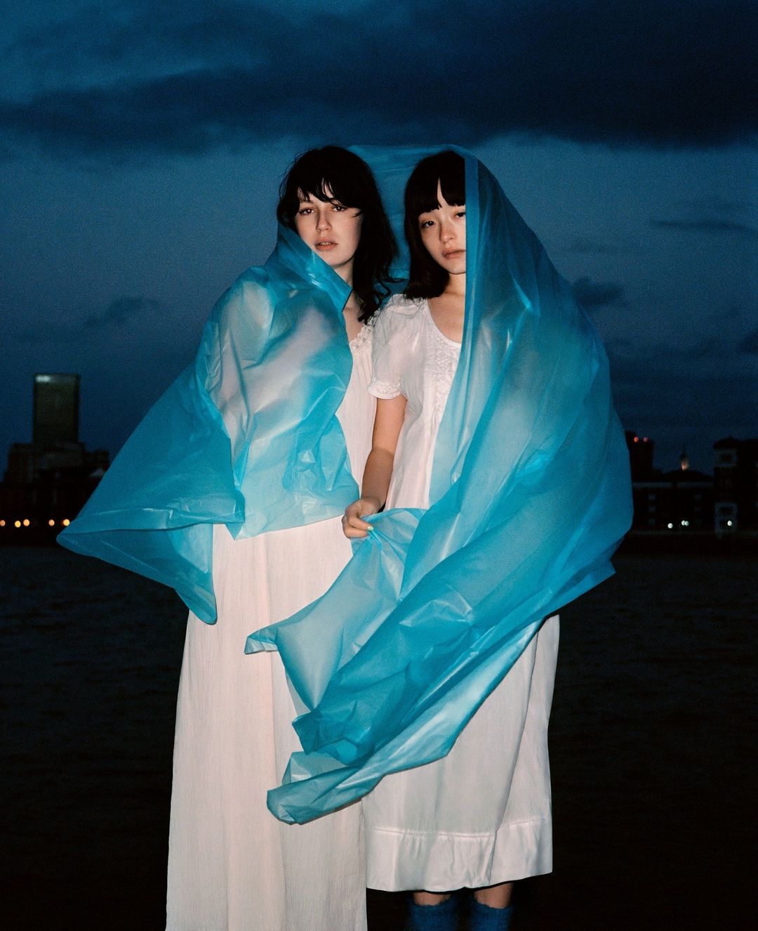 Two young women in white dresses and transparent turquoise wraps pose outdoors against a twilight sky and city buildings.