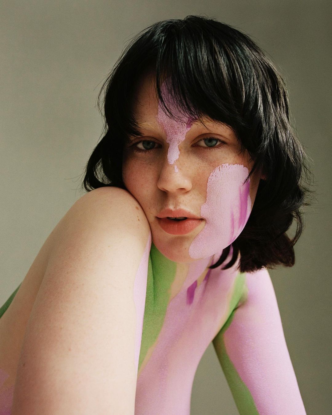 Close-up portrait of a young adult woman with short dark hair, light skin, and pink and green body paint on a light gray background.