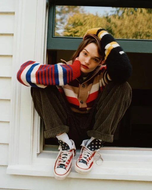 Stylish woman in striped cardigan and American flag sneakers sits in window, looking at camera, with natural light and blurred background.