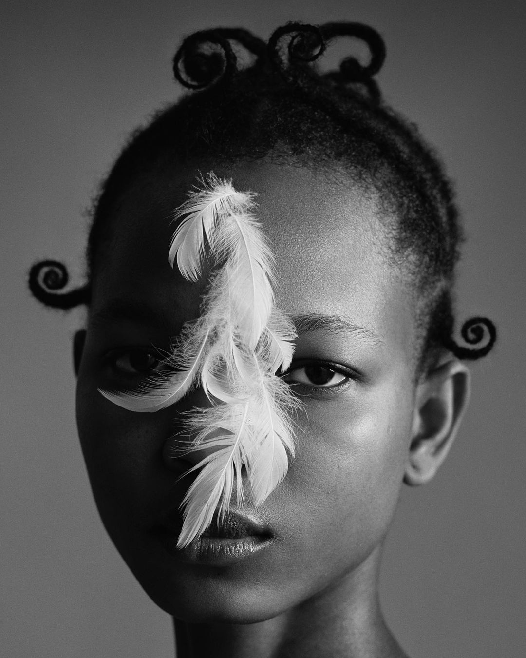 Close-up black and white portrait of a woman with curly hair, accentuated by white feathers covering one eye in a studio setting.