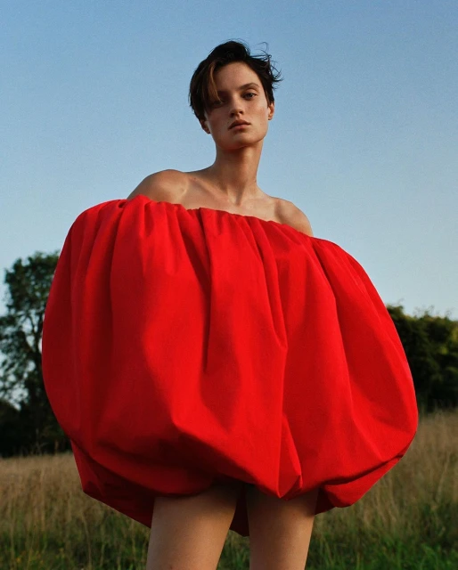 Portrait of a young woman in a voluminous red off-the-shoulder dress, posing in a grassy field against a blue sky and scattered trees.