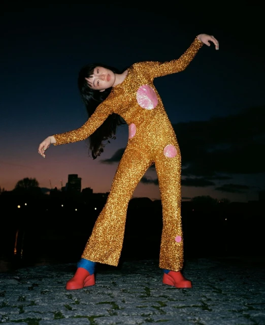 Woman in shimmering gold jumpsuit with pink circles poses on cobblestone street at night with city buildings in the background.