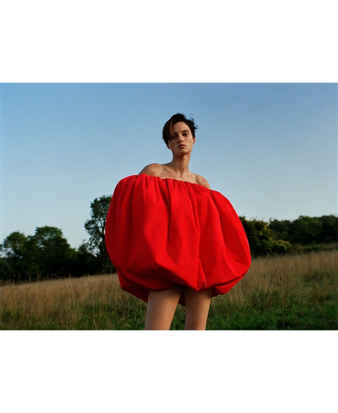 Model wearing a voluminous red off-the-shoulder dress standing in an outdoor grassy field under a clear blue sky