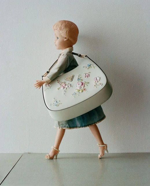 Fashion photography featuring a doll carrying a Prada handbag, wearing a denim skirt and heels, against a neutral background.
