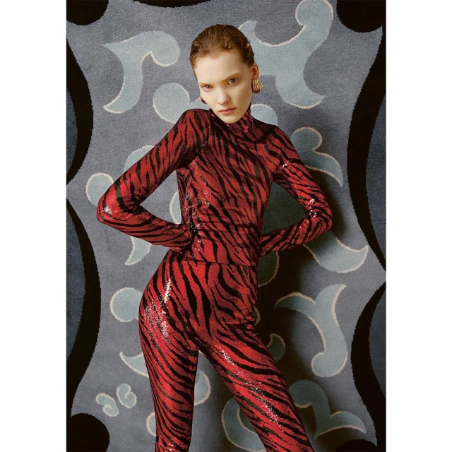 A fashionable woman in a red and black tiger-striped outfit stands in front of a patterned blue and gray backdrop. Medium shot.