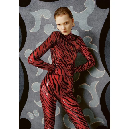 A fashionable woman in a red and black tiger-striped outfit stands in front of a patterned blue and gray backdrop. Medium shot.