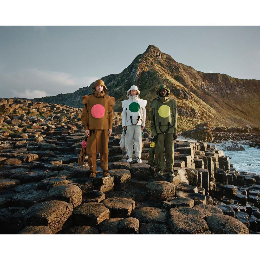 Three people in color-blocked outfits standing on hexagonal rock formations with a mountain and the ocean in the background.