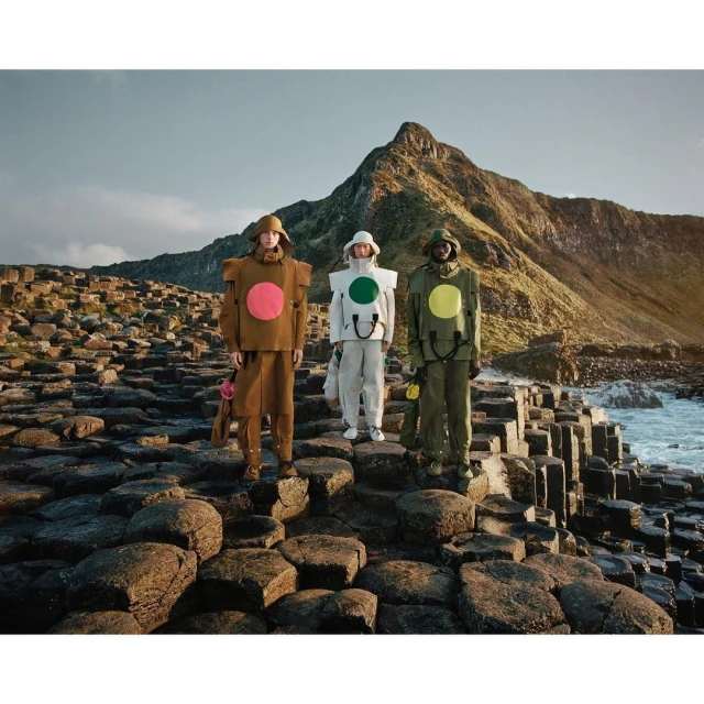 Three people in color-blocked outfits standing on hexagonal rock formations with a mountain and the ocean in the background.