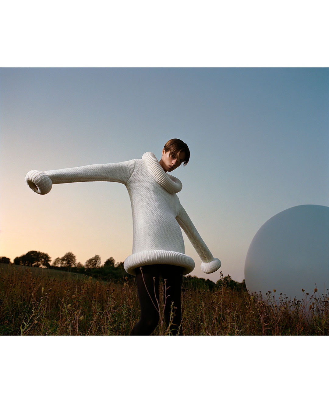 A person in a large, white knit sweater with oversized sleeves stands in a grassy field under a blue sky with a white sphere nearby.