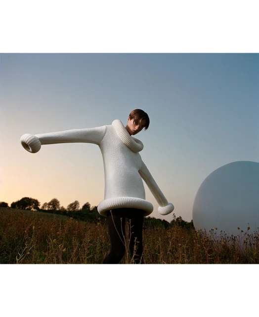 A person in a large, white knit sweater with oversized sleeves stands in a grassy field under a blue sky with a white sphere nearby.