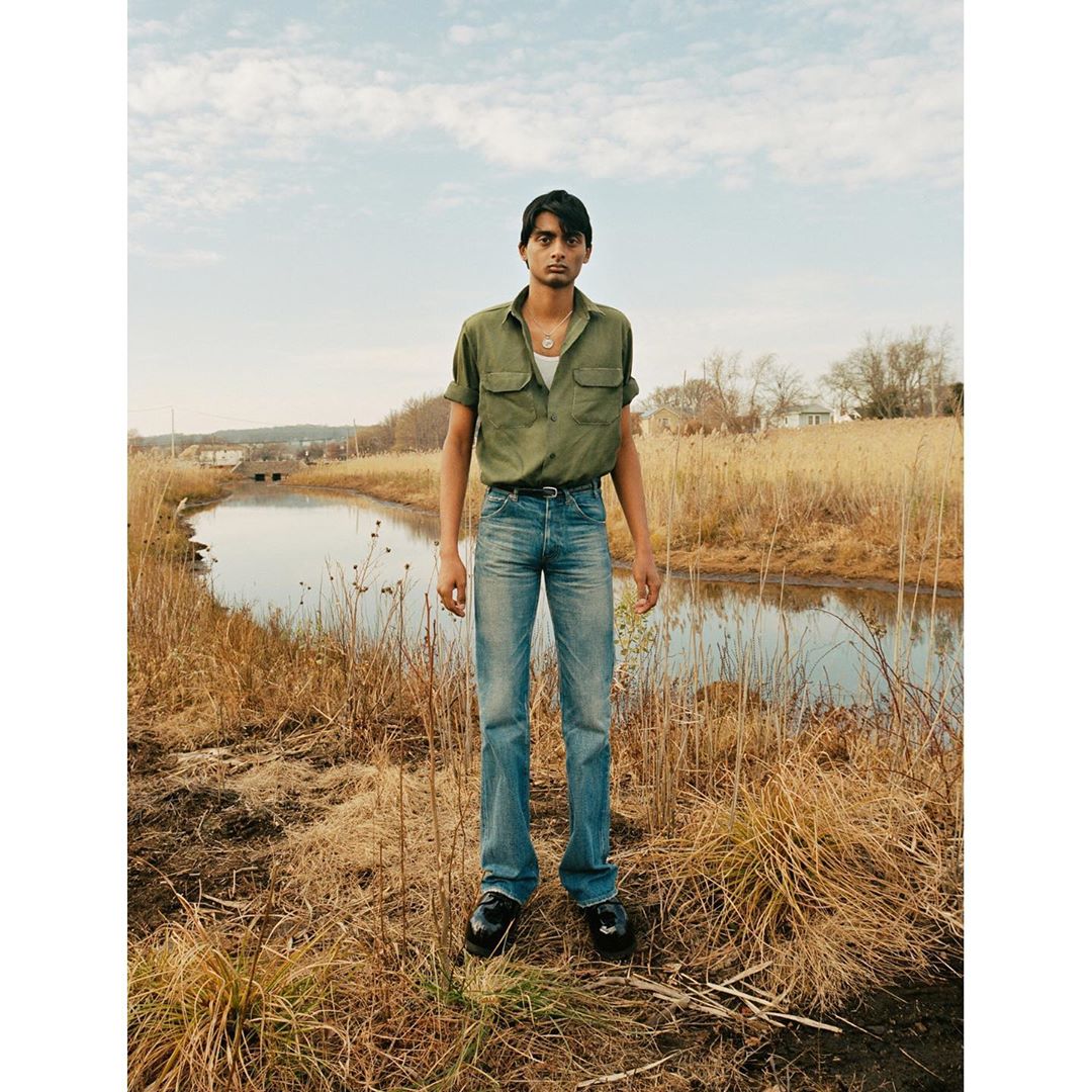 A young south asian man wearing a green short-sleeve shirt and blue jeans standing outdoors near a riverbank.
