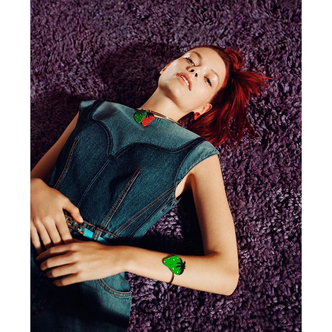 Adult woman with auburn hair lies on a purple shag rug wearing a denim vest, belt with turquoise accents, and strawberry jewelry.