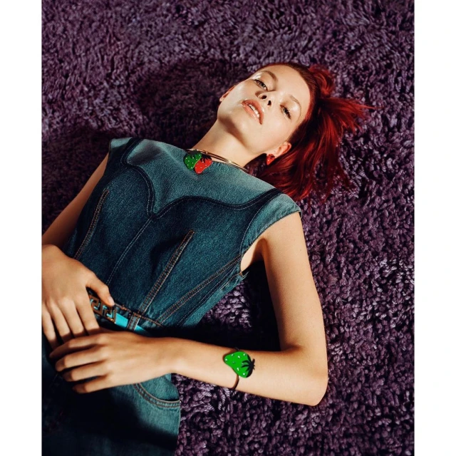 Adult woman with auburn hair lies on a purple shag rug wearing a denim vest, belt with turquoise accents, and strawberry jewelry.