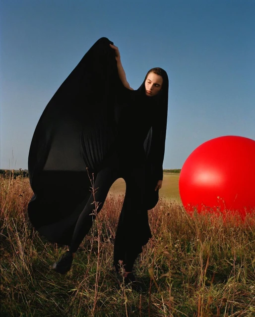 Woman in a black draped costume stands in a grassy field with a large red ball, under a clear blue sky, creating a surreal outdoor scene.