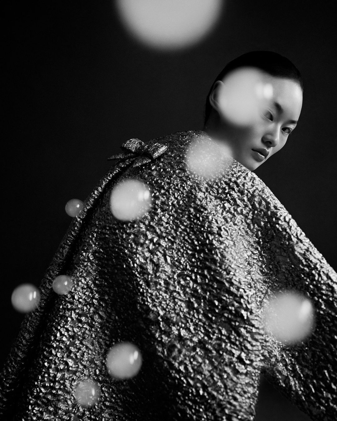 Monochrome portrait of a stylish Asian woman with short hair, posing in a reflective textured coat against a dark backdrop, studio lighting.